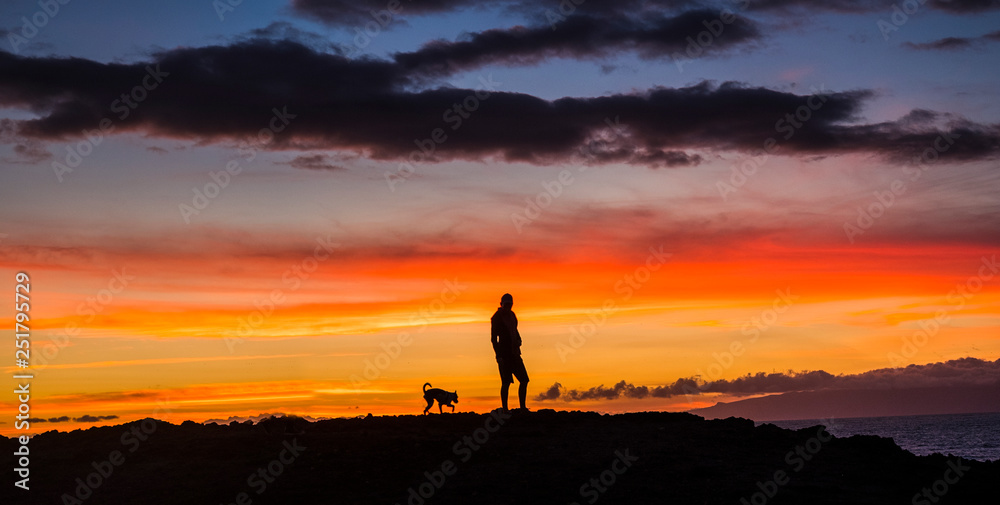 Man standing in the sunset with best friend dog animal with him - friendship and romantic outdoor leisure activity for people - colors and red clouds - vacation enjoying life
