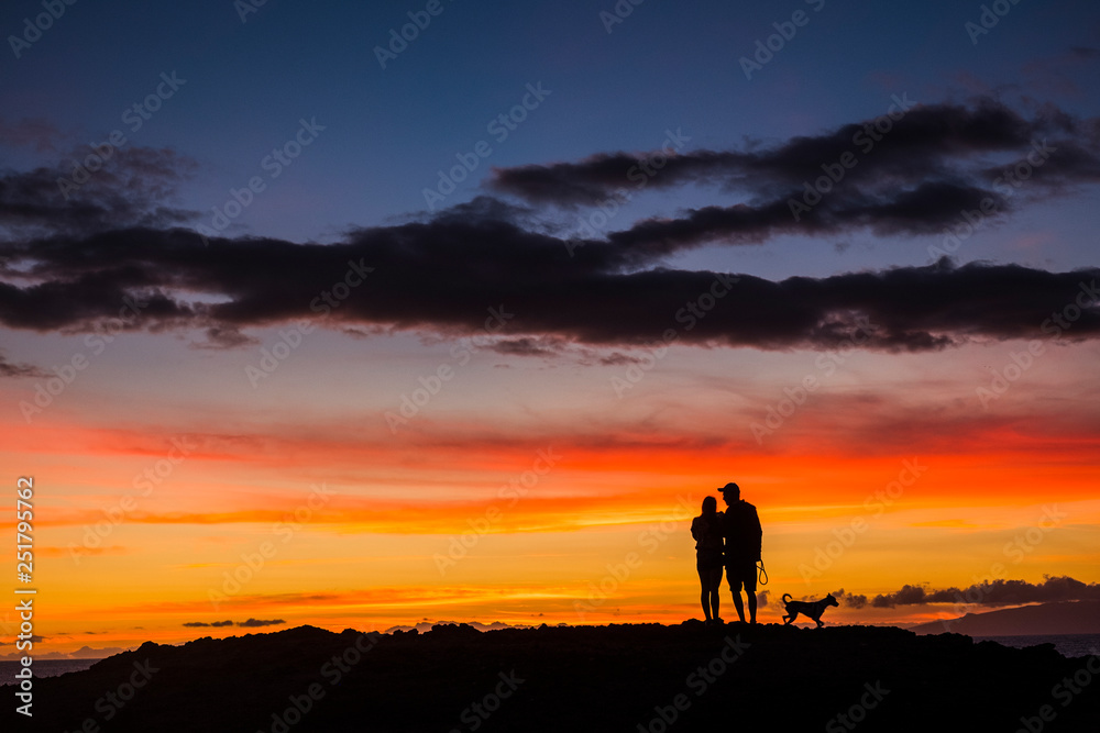 Fototapeta premium Romantic couple wioth dog enjoying and looking the sunset with red and orange coloured clouds in the sky - wanderlust and travel concept for happy people - dawn and colors background concept