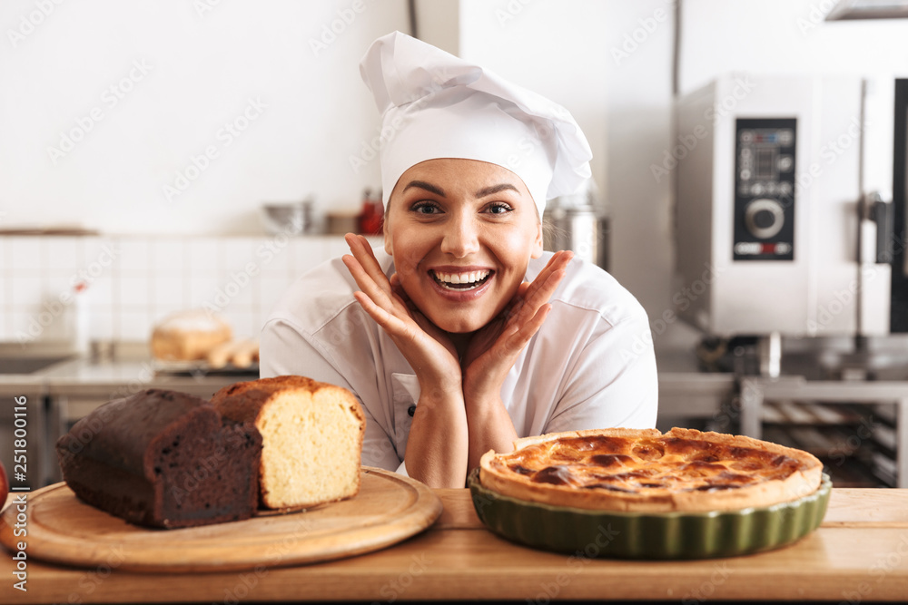 Image of adult woman chef wearing white uniform, posing in kitchen at the cafe with baked goods Image of adult woman chef wearing white uniform, posing in kitchen at the cafe with baked goods