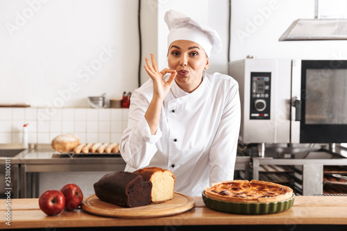 Image of beautiful woman chef wearing white uniform, posing in kitchen at the cafe with baked goods