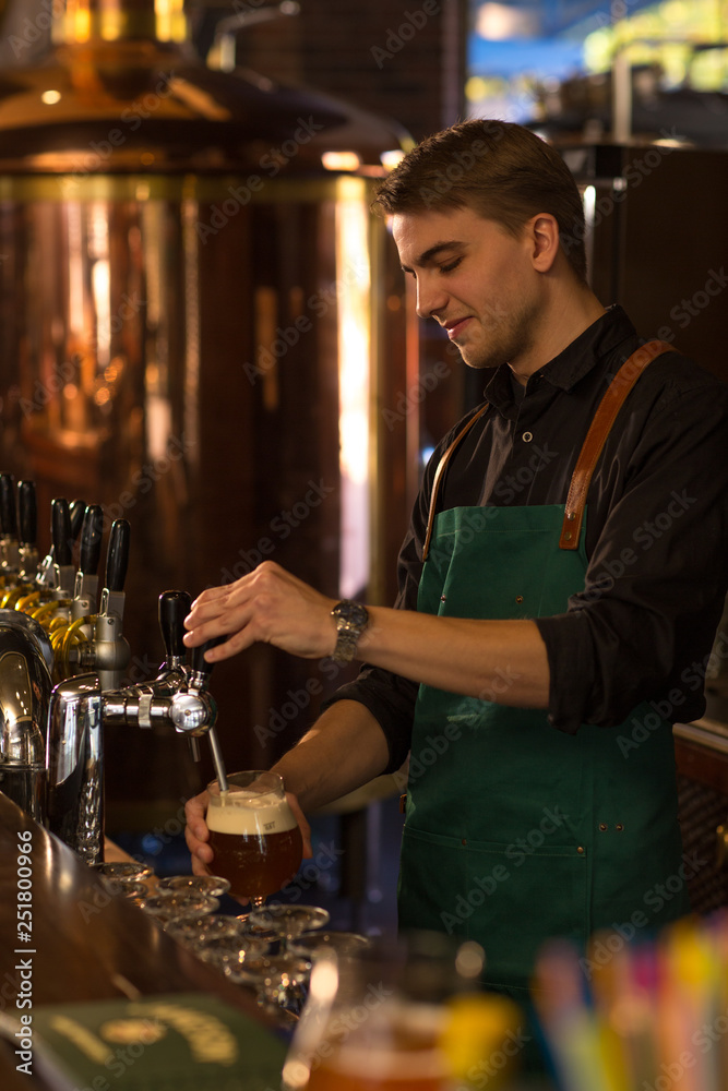 Cheerful barman pouring beer, looking down. Man wearing in black shirt and dark green apron while working in brewery. Big bronzed kettle or reservoir in beer house behind.
