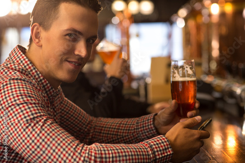 Client of beer house posing with glass of beer in hand. Handsome man looking at camera, smiling, holding mobile phone. Man wearing in checked shirt resting, enjoying alcohol beverages.