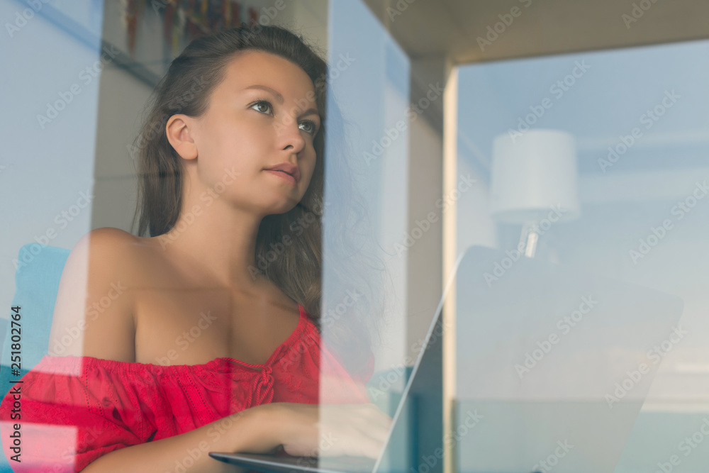 Attractive young woman with a laptop sits on a sofa in a cafe behind the glass of window. Lifestyle concept. Attractive young woman with a laptop sits on a sofa in a cafe behind the glass of window. Lifestyle concept.