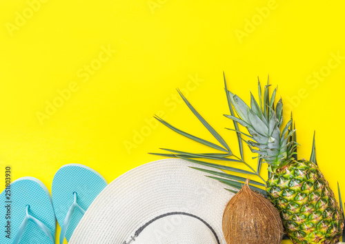 Ripe pineapple coconut on green palm leaf white straw hat blue slippers on bright yellow solid background. Summer vacation fun tropical fruits beach party concept. Trendy flat lay