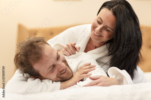 Happy man lying on bed while beautiful wife lying on his back and looking at him. Cheerful couple in white robes laughing and relaxing in morning in hotel together. Concept of love and happiness.
