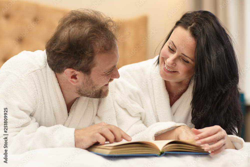 Cheerful couple in white robes laying on bed at home and discussing interesting story. Satisfied wife and husband reading new book together and enjoying resting. Concept of time spending. Cheerful couple in white robes laying on bed at home and discussing interesting story. Satisfied wife and husband reading new book together and enjoying resting. Concept of time spending.