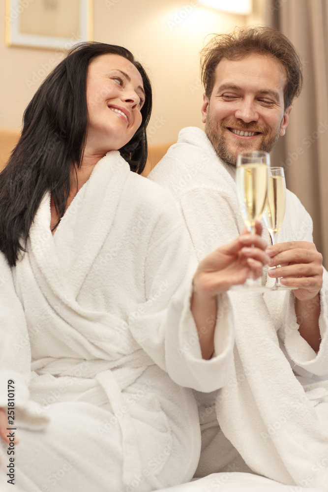 Wonderful couple sitting back to back on bed and drinking champagne together. Attractive brunette and bearded man cheering, looking and each other and smiling. Concept of romantic atmosphere. Wonderful couple sitting back to back on bed and drinking champagne together. Attractive brunette and bearded man cheering, looking and each other and smiling. Concept of romantic atmosphere.