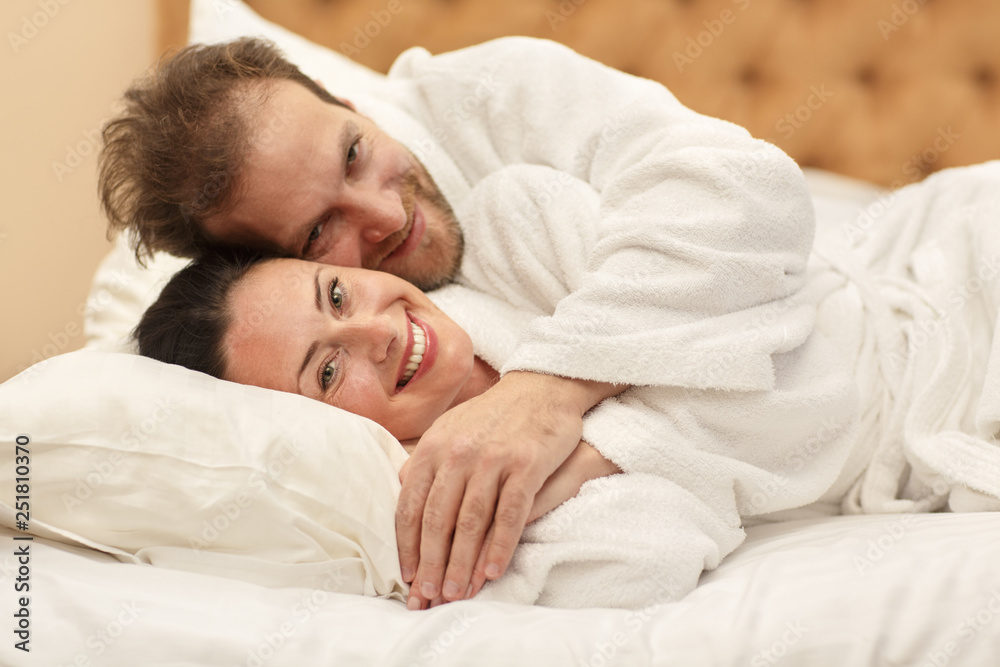 Lovely adults lying on bed, looking at camera and smiling. Bearded husband and attractive wife in white robes hugging and getting up together in morning. Concept of good morning and love. Lovely adults lying on bed, looking at camera and smiling. Bearded husband and attractive wife in white robes hugging and getting up together in morning. Concept of good morning and love.