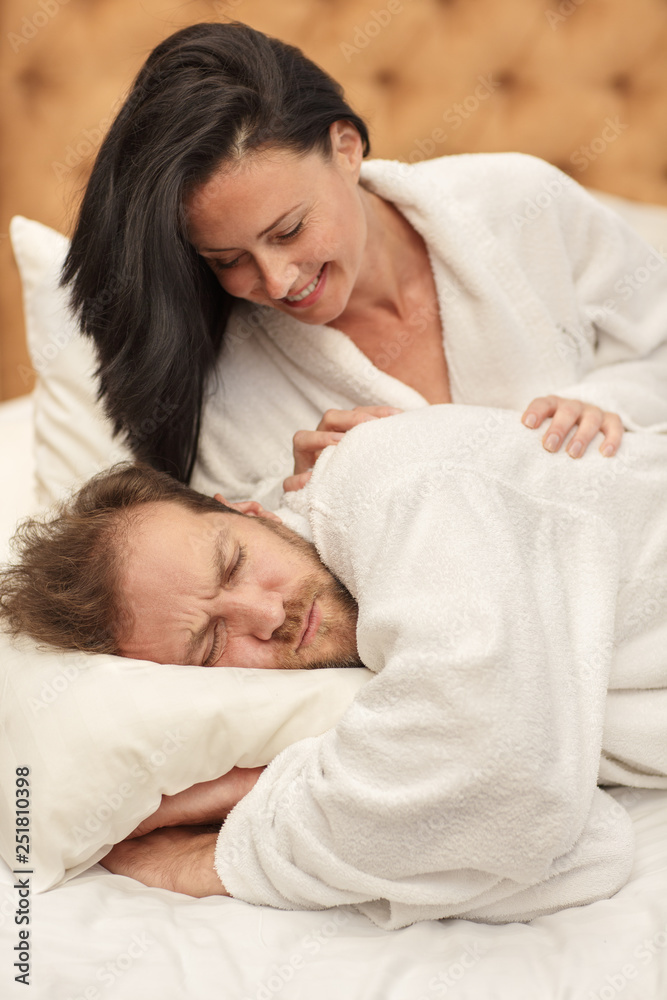 Beautiful smiling woman in white robe awakening bearded male lying in bed. Tired husband embracing pillow and soundly sleeping in morning. Concept of getting up and dreaming. Beautiful smiling woman in white robe awakening bearded male lying in bed. Tired husband embracing pillow and soundly sleeping in morning. Concept of getting up and dreaming.