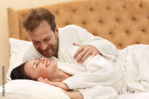 Happy couple lying together on bed, hugging, looking at each other and smiling. Female and male in white robes enjoying good morning and time together. Concept of love and happiness.