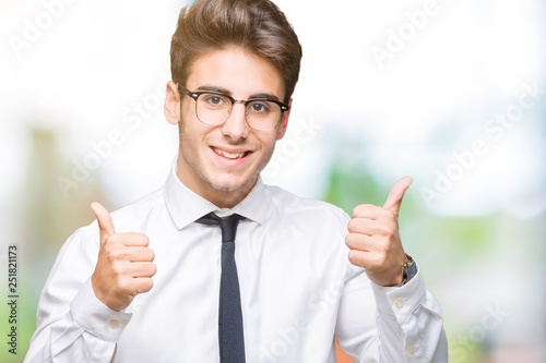 Young business man wearing glasses over isolated background success sign doing positive gesture with hand, thumbs up smiling and happy. Looking at the camera with cheerful expression, winner gesture.