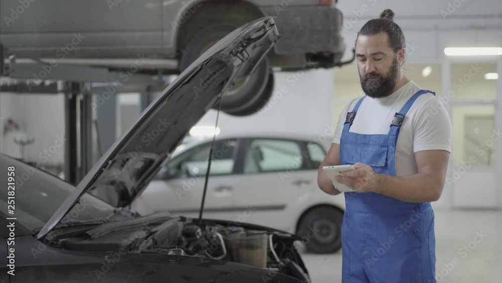 Adult mechanic standing near the car with an open hood and verifies information on his phone. Bearded man in uniform fixing automobile in car repair station Adult mechanic standing near the car with an open hood and verifies information on his phone. Bearded man in uniform fixing automobile in car repair station