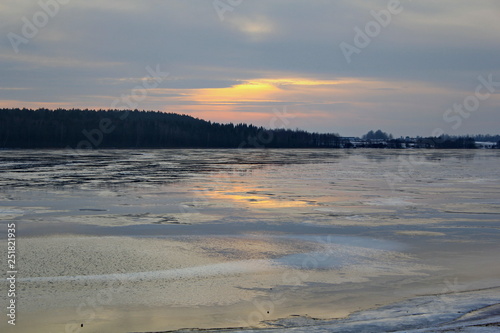 The rays of the sunset sun breaking through the gray clouds and reflected in the surface of the lake with the remains of ice and dark banks covered with forest