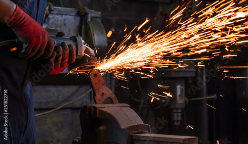 Body shot of mechanical engineer worker wearing safety gloves and operating a angle grinder on his workbench to metal held in an iron vice inside a workshop with flash sparks