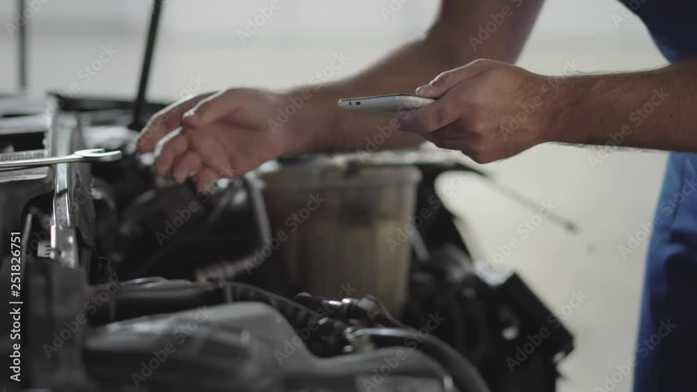 Hands of auto master working with the car with an open hood and verifies information on his phone. Skillful man in uniform fixing automobile in car repair station Hands of auto master working with the car with an open hood and verifies information on his phone. Skillful man in uniform fixing automobile in car repair station
