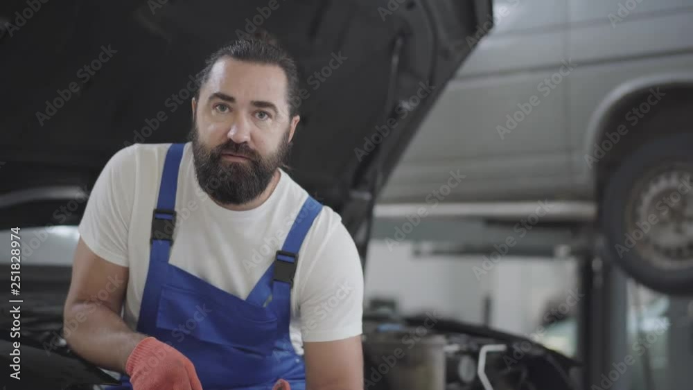 Portrait of handsome bearded mechanic posing with spanner sitting on the hood of a broken car in a car repair shop. Portrait of handsome bearded mechanic posing with spanner sitting on the hood of a broken car in a car repair shop.