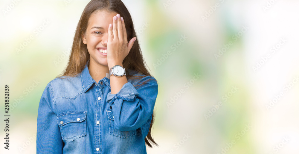 Young beautiful brunette woman wearing blue denim shirt over isolated background covering one eye with hand with confident smile on face and surprise emotion. Young beautiful brunette woman wearing blue denim shirt over isolated background covering one eye with hand with confident smile on face and surprise emotion.