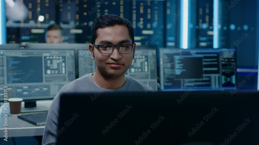 Portrait of a Smart and Handsome Software Developer Wearing Glasses Working on a Personal Computer, Smiles at the Camera. In the Background Unfocused Personal Computers with Screens in Technical Room Portrait of a Smart and Handsome Software Developer Wearing Glasses Working on a Personal Computer, Smiles at the Camera. In the Background Unfocused Personal Computers with Screens in Technical Room