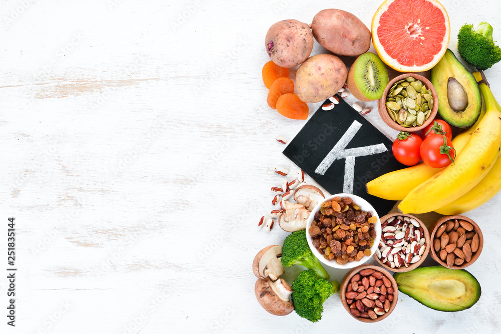 Foods containing natural potassium. K: Potatoes, mushrooms, banana, tomatoes, nuts, beans, broccoli, avocados. Top view. On a white wooden background. Foods containing natural potassium. K: Potatoes, mushrooms, banana, tomatoes, nuts, beans, broccoli, avocados. Top view. On a white wooden background.