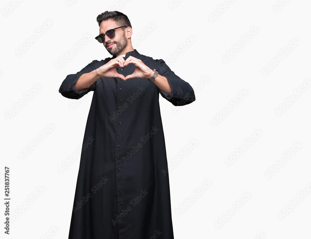 Young Christian priest wearing sunglasses over isolated background smiling in love showing heart symbol and shape with hands. Romantic concept.