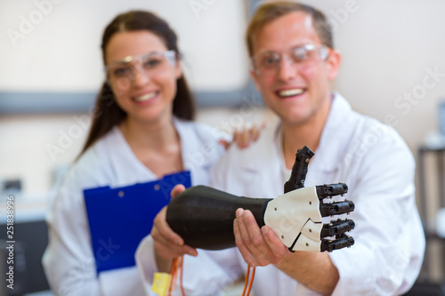 Attractive scientist holding plastic hand and gesturing thumb up. Male engineer, wearing in white uniform, testing invention, smiling and looking at camera while her colleague standing near.