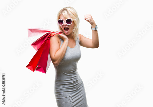 Young beautiful blonde woman shopping holding shopping bags on sales over isolated background annoyed and frustrated shouting with anger, crazy and yelling with raised hand, anger concept
