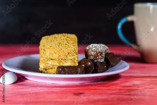 Layered sponge cake, chocolate sweets and cup of coffee on wooden background.