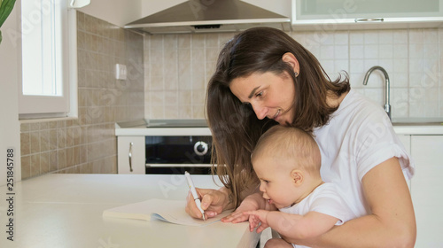 Young mother writes in a notebook and plans with baby in the kitchen at home