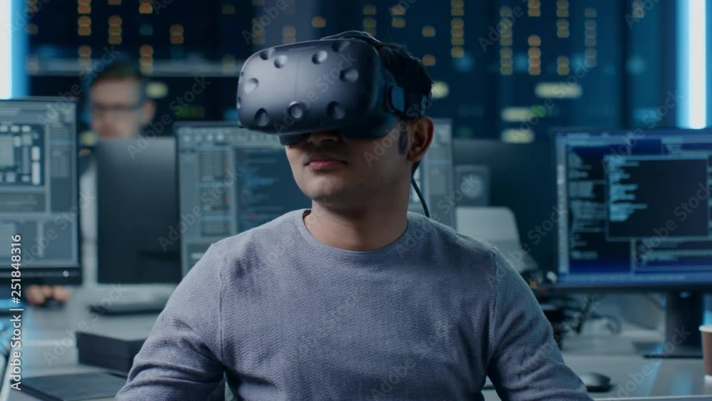 Software Delevoper Wearing Virtual Reality Headset Gesturing with His Head, Developing and Programming VR Game or Application. In Background Technology Developing Studio with Computers and Monitors Software Delevoper Wearing Virtual Reality Headset Gesturing with His Head, Developing and Programming VR Game or Application. In Background Technology Developing Studio with Computers and Monitors