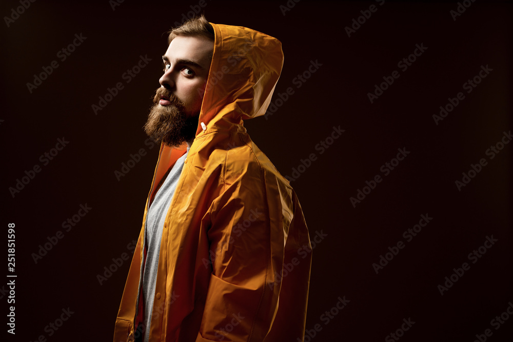Guy with a beard and mustache dressed in a gray t-shirt and yellow jacket with a hood is standing on a brown background in the studio