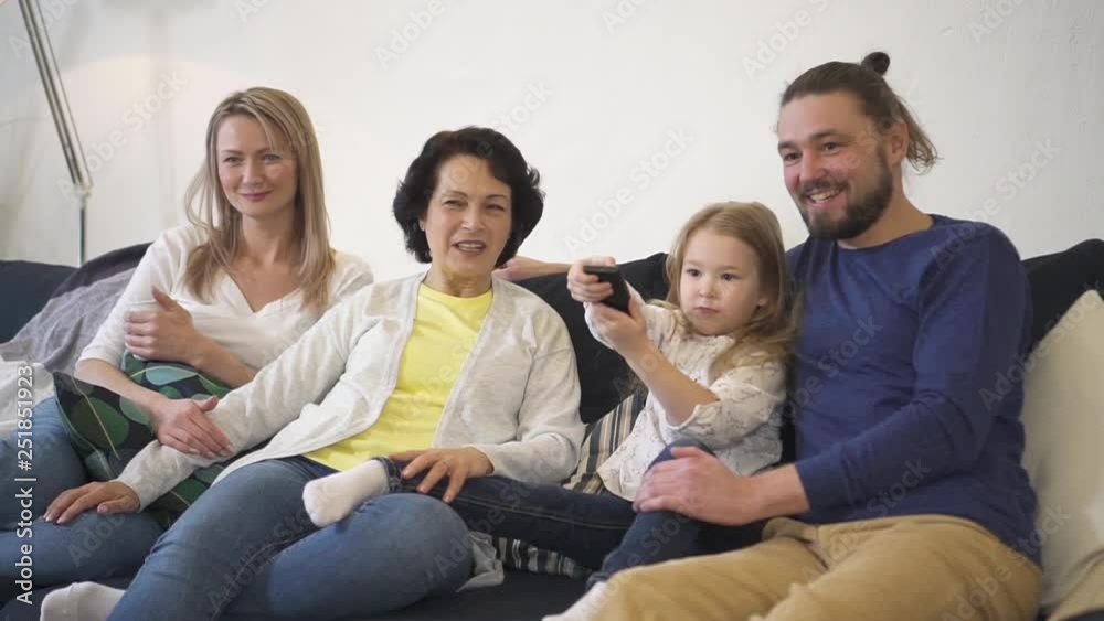 Whole happy family is sitting on sofa and relaxing by choosing and watching tv. Father is holding little daughter while senior mature grandmother smiling at girl. Entertainment television technology. Whole happy family is sitting on sofa and relaxing by choosing and watching tv. Father is holding little daughter while senior mature grandmother smiling at girl. Entertainment television technology.