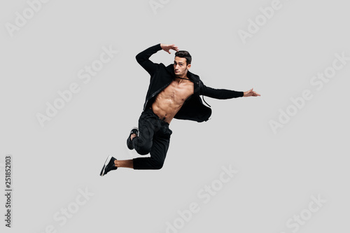 Handsome young dancer of street dancing dressed in black pants and a sweatshirt on a naked torso jumps on a white background
