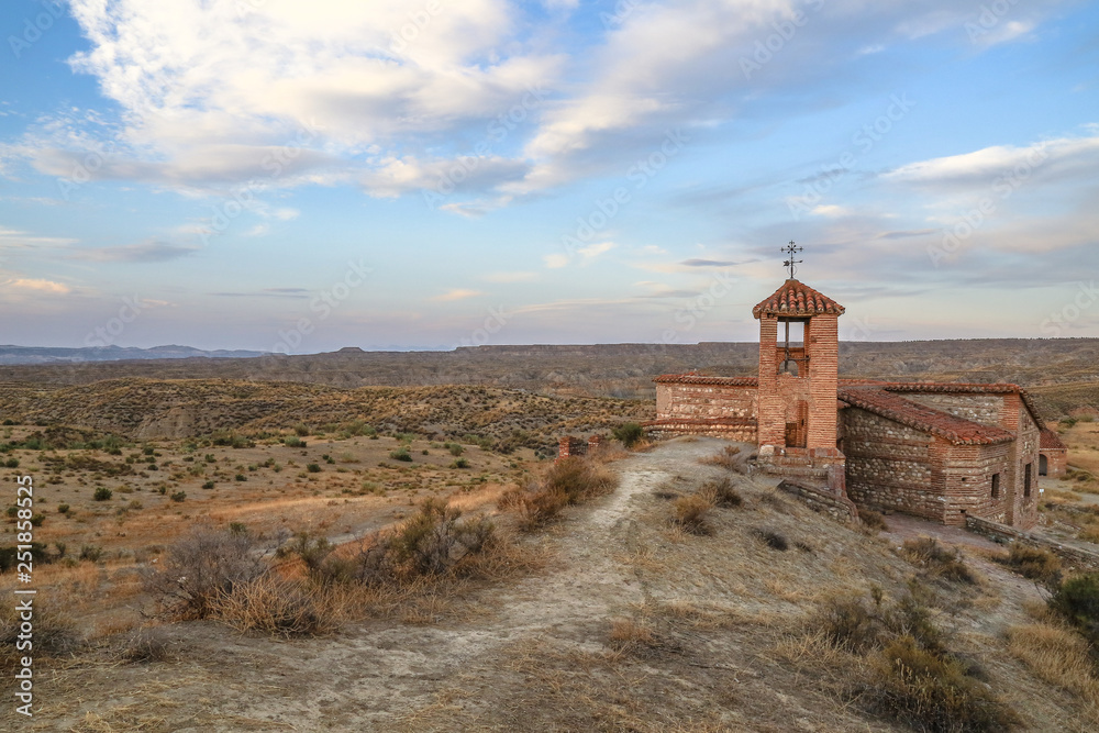 old abandoned red coloured church in the middle of nowhere surrounded by dry steppe grassland desert under a blue sky with clouds