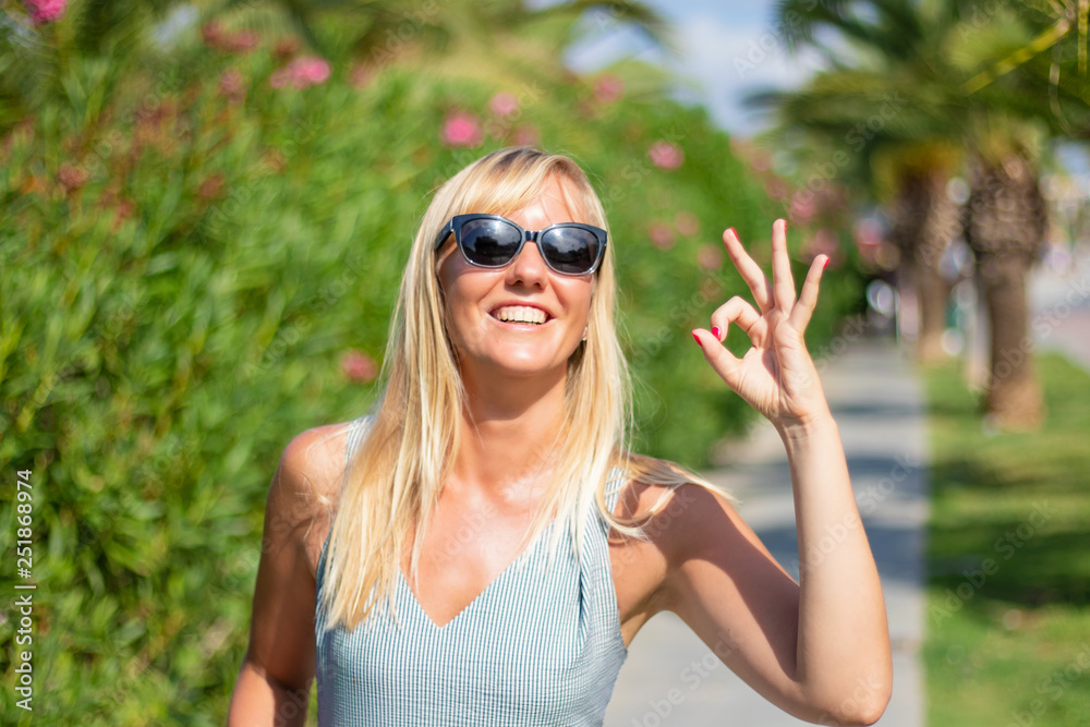 Portrait of beautiful young happy smiling blonde woman with okay gesture. Blooming spring garden background. Portrait of beautiful young happy smiling blonde woman with okay gesture. Blooming spring garden background.