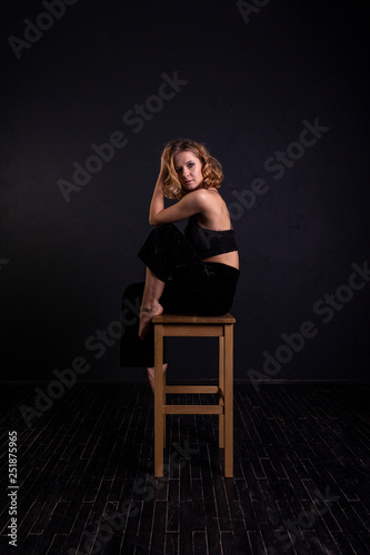 A pretty woman with a good figure is standing in a bra and black pants near the bar stool in the room.