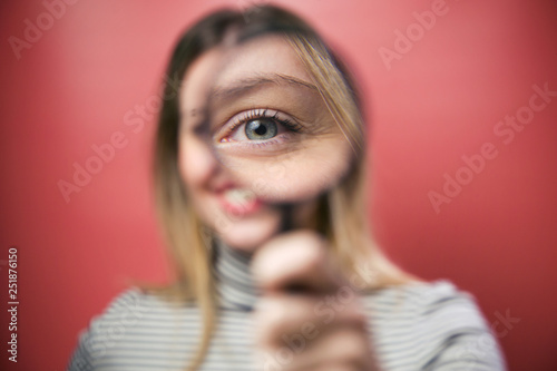 Beautiful young woman looking through magnifying glass at the camera over pink background.