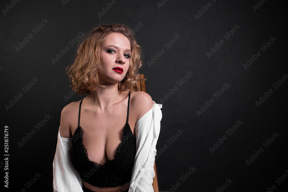 Beautiful, sexy and fashionable blonde girl with red lips in white shirt, black bra and black leggings sits on a wooden chair on black background Beautiful, sexy and fashionable blonde girl with red lips in white shirt, black bra and black leggings sits on a wooden chair on black background
