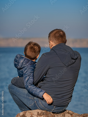 Cute European boy and his dad are sitting on the stone at the bank of the lake. They are looking ahead and enjoying fresh air and serenity around...