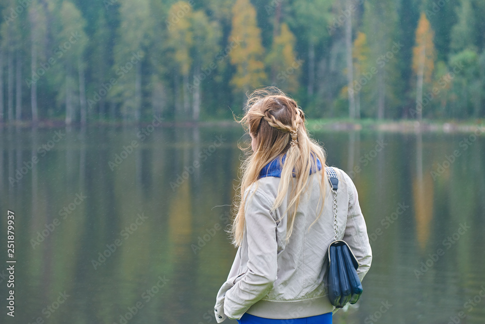 Fototapeta premium The portrait of a beautiful young woman with long blonde hair on the wooden quay against the forest and lake.