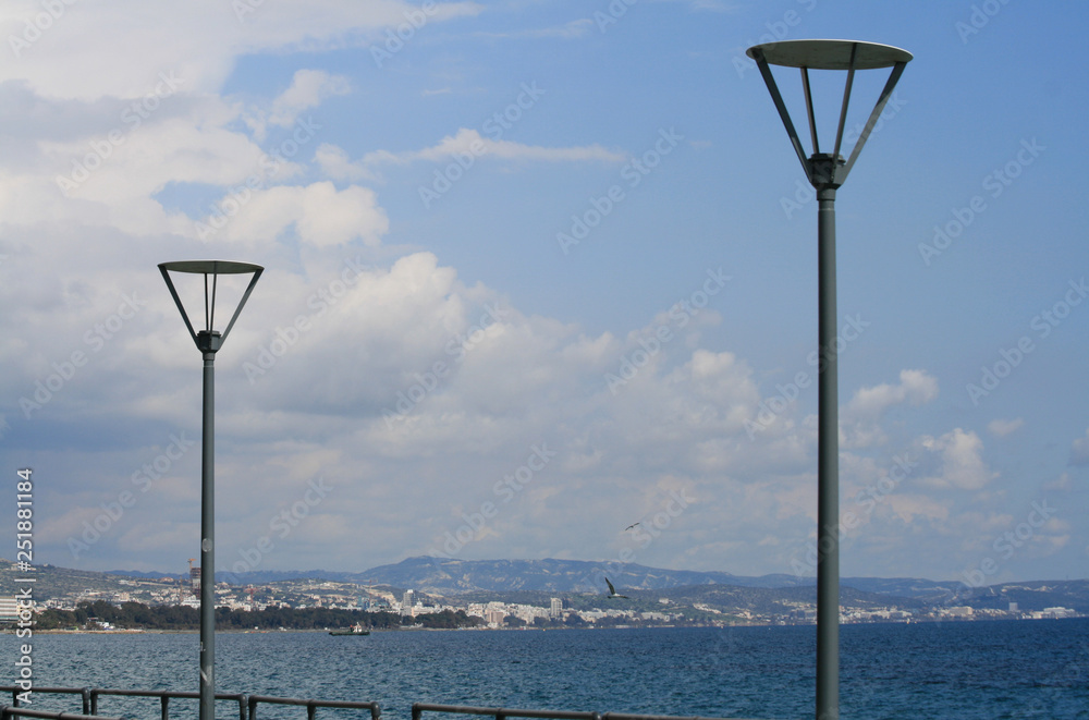 Two metal street lamps on a pier in front of the blue sea, sky with white clouds and a view of a coastline
