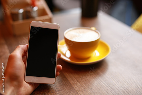 Male hand holding smartphone in cafe, yellow cup with latte on background