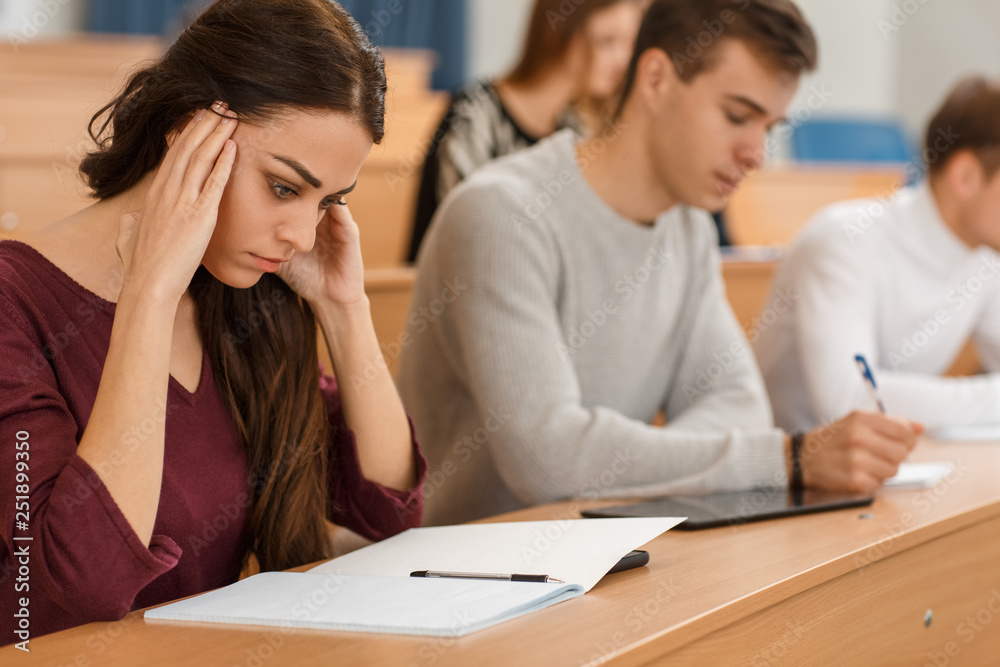 Serious female student sitting at table in lecture hall, keeping hands near head and thinking over difficult test. Concentrated girl looking down at notebook at university. Concept of studying. Serious female student sitting at table in lecture hall, keeping hands near head and thinking over difficult test. Concentrated girl looking down at notebook at university. Concept of studying.