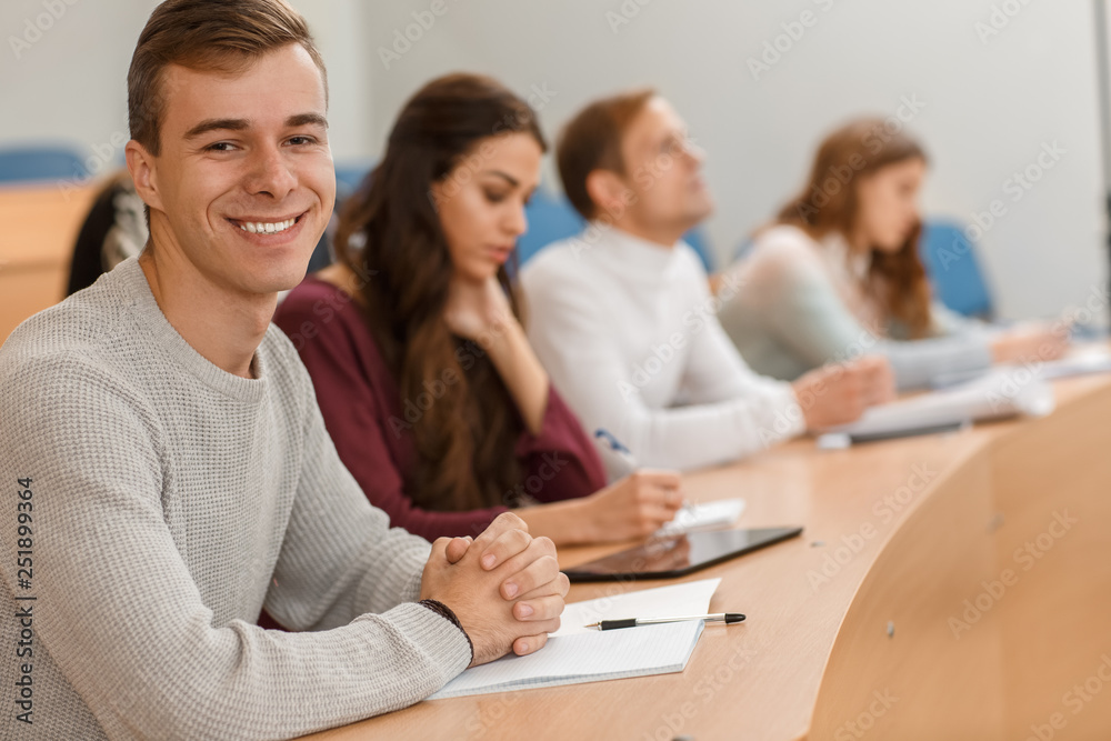 Handsome male student sitting at table in lecture hall, looking at camera and smiling. Cheerful man writing notes and enjoying studying process at university. Concept of interesting education. Handsome male student sitting at table in lecture hall, looking at camera and smiling. Cheerful man writing notes and enjoying studying process at university. Concept of interesting education.