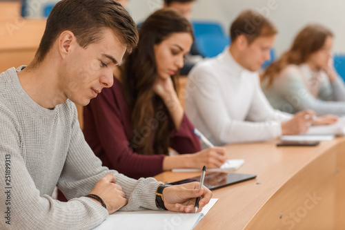 Cunning male student in white sweater sitting at table, smiling and copying notes at university. On background preyyt brunette girl learning and getting high education. Concept of in studying.