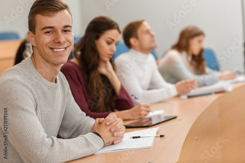 Handsome male student sitting at table in lecture hall, looking at camera and smiling. Cheerful man writing notes and enjoying studying process at university. Concept of interesting education.