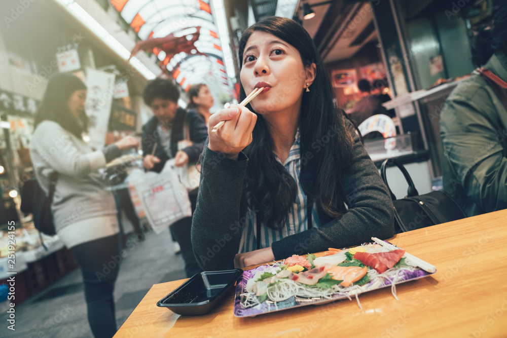 vintage photo of joyful japanese woman eat fresh and raw food tuna sashimi in plate for sale. young happy girl sitting at vendor having snack with chopsticks in Kuromon street market Osaka Japan.
