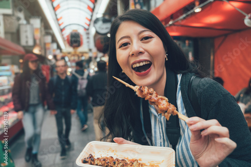 local food festival japanese market concept. female traveler visit kuromon ichiba osaka trying delicious street food of barbecue chicken. happy lady showing tasty snack in teeming place. photo