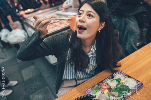 female asian visitors visit Kuromon Ichiba Market Japan. girl eating fresh raw fish seafood in popular area known by locals as Osaka kitchen. woman traveler sitting at vendor having delicious meal photo