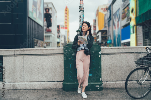 full length of young girl traveler with slr camera holding paper map searching right way to destination standing on bridge walk way with bicycle near by. woman relaxing tourism osaka outdoor. photo