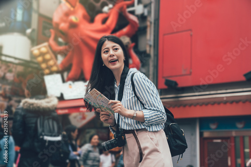 vintage style photo of smiling gril backpack photographer standing in front of takoyaki food vendor shop with huge octopus animal advertisement hanging on wall in background. lady cheerful with map photo